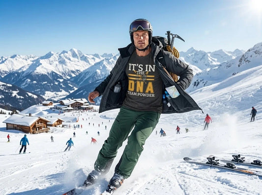 Its in the DNA A man skiing down a snowy mountain slope wearing a dark gray graphic t-shirt with 'IT'S IN THE DNA TEAM POWDER' printed on the front, green ski pants, and a black jacket, with mountains and other skiers in the background.