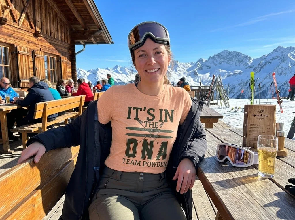 It's in the DNA A woman sitting at an outdoor wooden table on a ski resort deck, wearing a peach-colored graphic t-shirt that reads 'IT'S IN THE DNA TEAM POWDER'. She has ski goggles on her head and a black jacket over her shoulders, with a glass of beer and more goggles on the table in front of her.