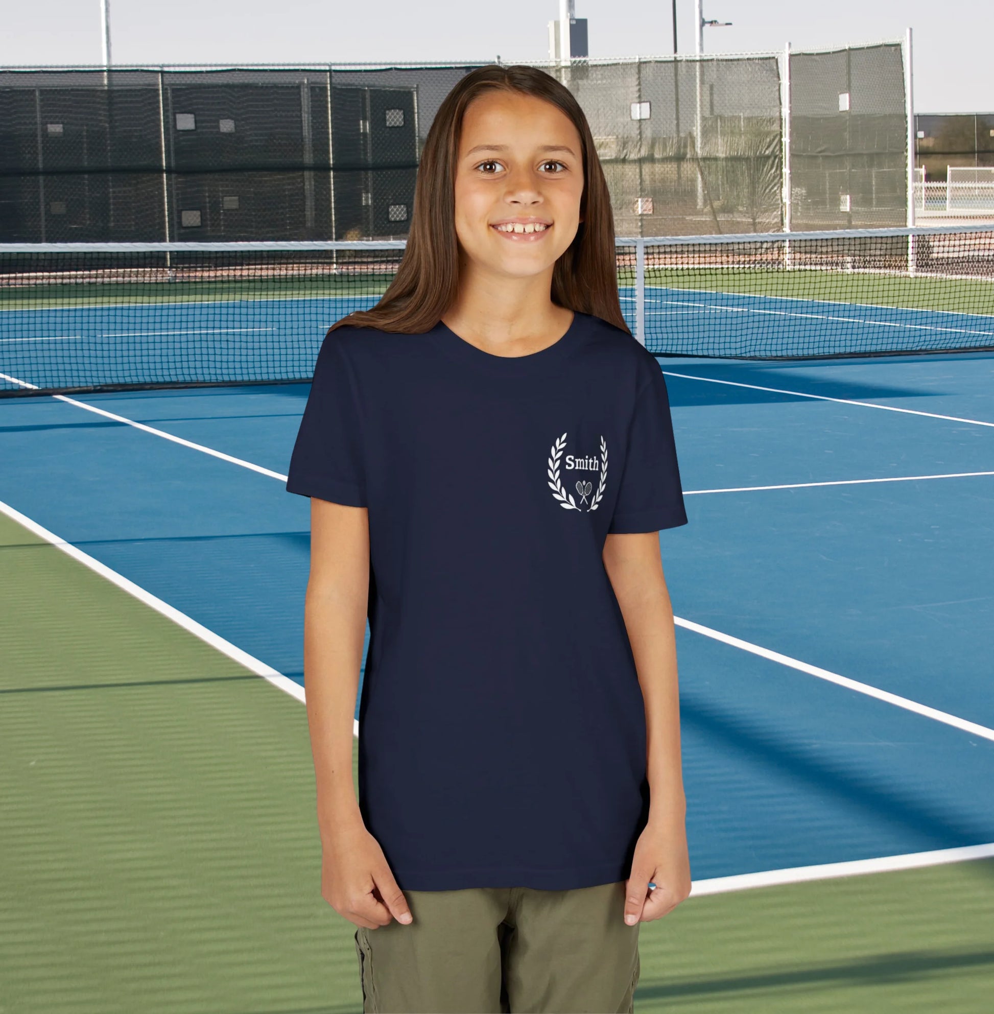 Junior tennis shirts Young girl standing on a tennis court wearing a navy blue t-shirt with a logo.