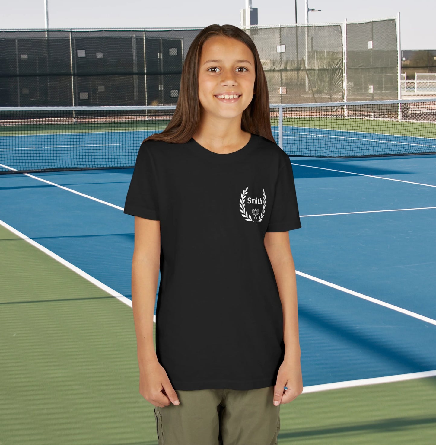Junior tennis shirts Young girl standing on a tennis court wearing a black t-shirt with a logo.