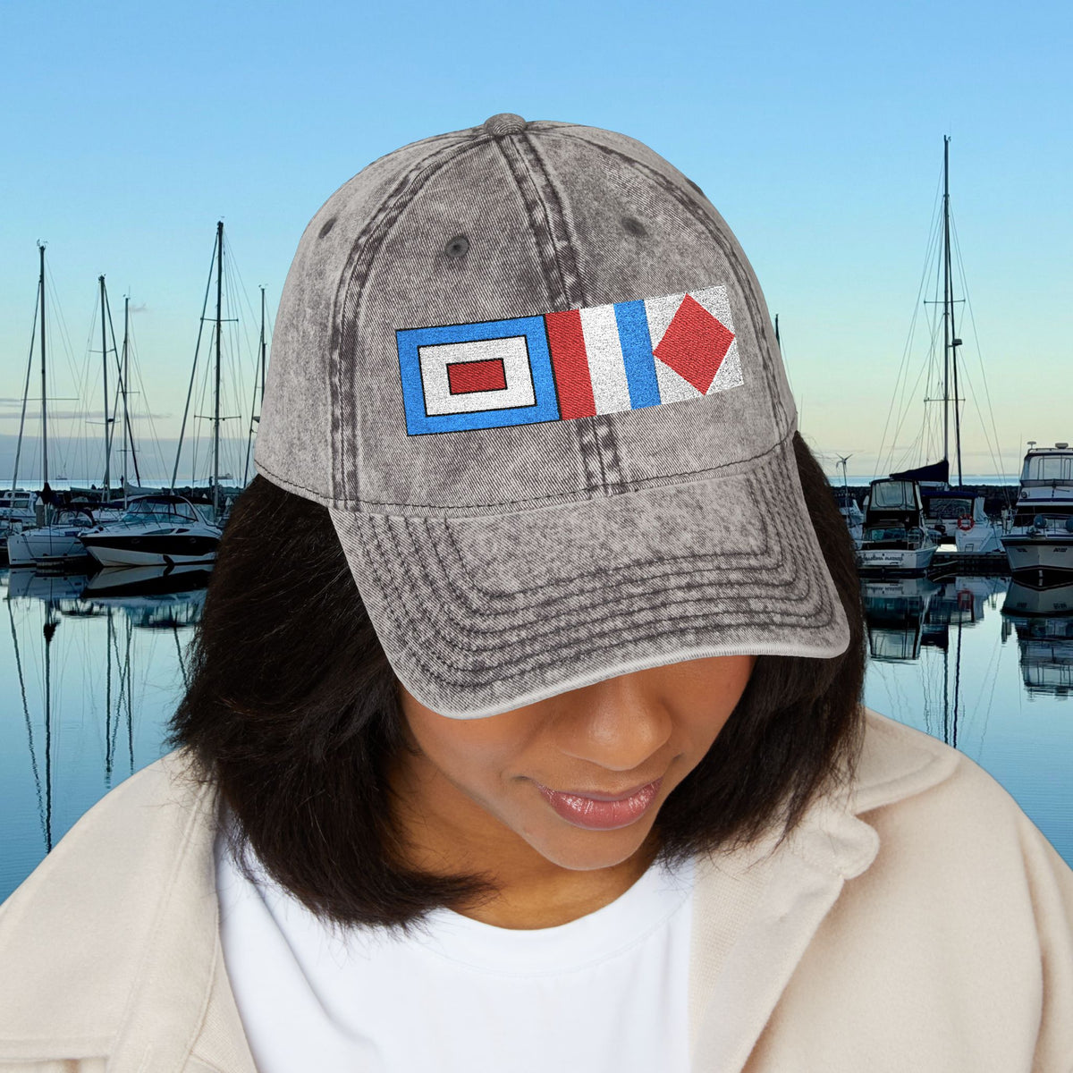 Nautical caps A person wearing a gray acid-wash baseball cap with embroidered nautical signal flags, set against a background of a calm harbor with boats and a blue sky.