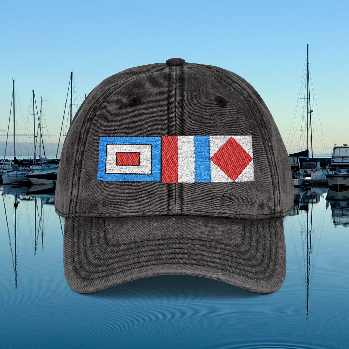 Nautical caps A dark gray washed baseball cap featuring three embroidered nautical signal flags on the front, set against a background of a calm harbor with boats and blue water.