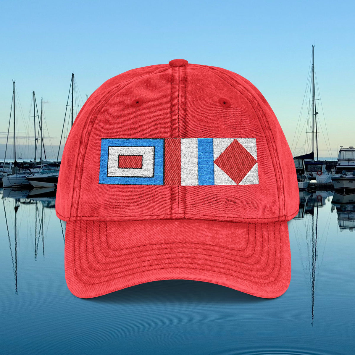 Nautical caps A red baseball cap with embroidered nautical signal flags on the front, set against a background of a calm harbor with boats and water under a blue sky.