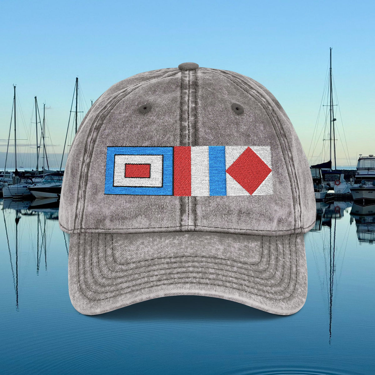 Nautical caps A gray washed baseball cap featuring a nautical signal flag patch on the front, set against a background of a calm harbor with boats and water under a blue sky.