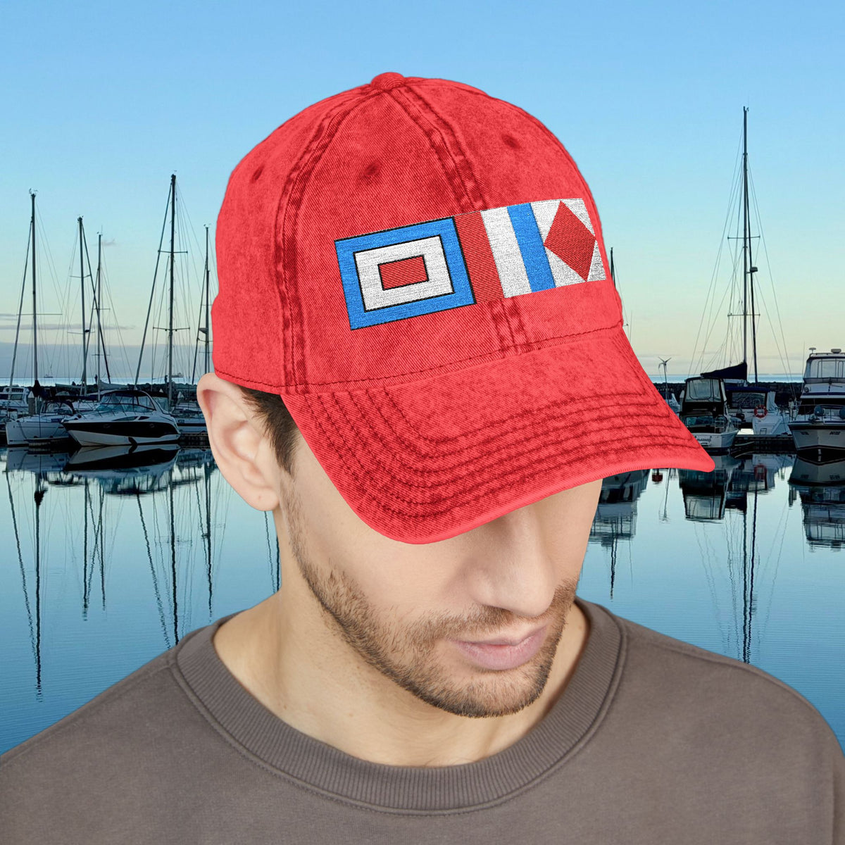 Nautical caps A man wearing a red baseball cap with two nautical signal flag patches, set against a background of a marina at dusk.