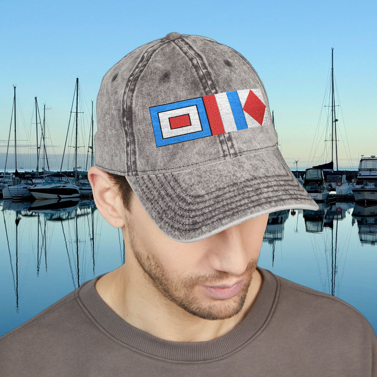 Nautical caps A man wearing a gray acid-wash baseball cap with a nautical flag patch on the front, set against a background of a marina at dusk.