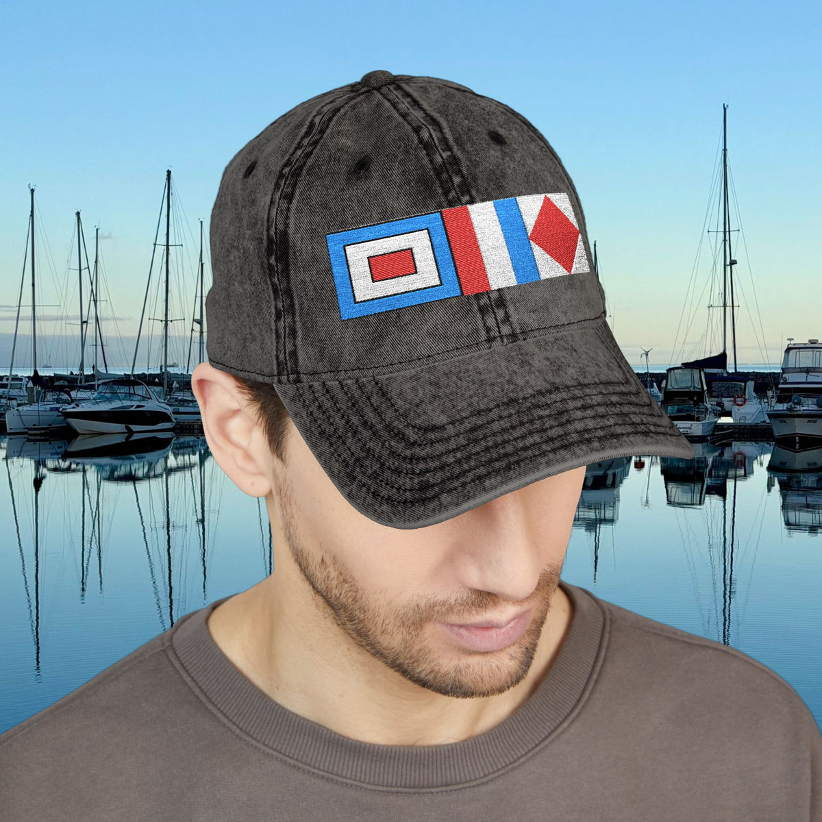 Nautical caps A man wearing a dark gray washed baseball cap with a nautical signal flag patch on the front, set against a background of a marina at dusk.