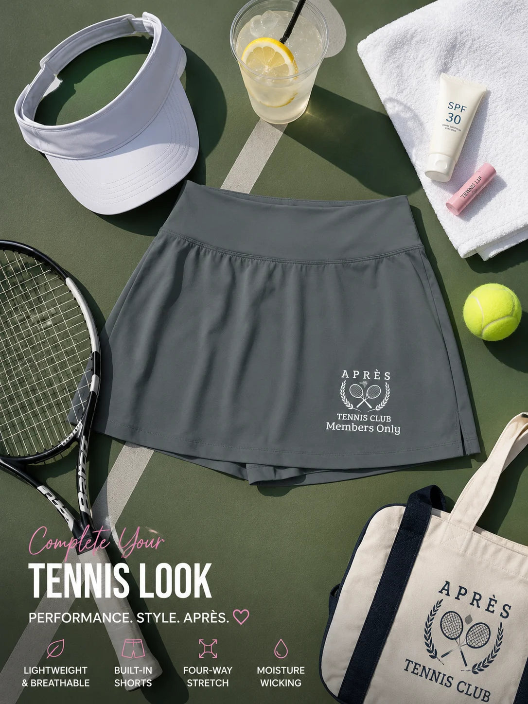 Personalized apres tennis club skort A dark gray athletic tennis skirt with a white printed logo is styled in a flat-lay on a green tennis court background with a tennis racket, visor, towel, sunscreen, tube, tennis ball, and tote bag.