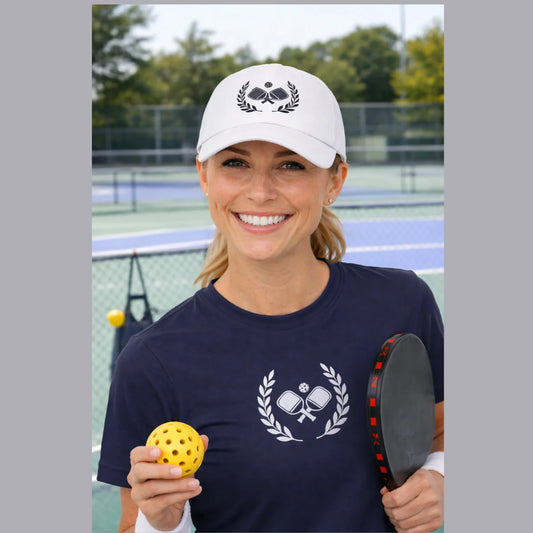 Pickleball  hats Woman on a pickleball court wearing a white cap and navy shirt with pickleball designs, holding a yellow ball and paddle.