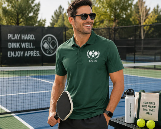 Pickleball Polos A man wearing a dark green polo shirt with a white embroidered logo and the name 'SMITH' stands on an outdoor pickleball court holding a paddle. A branded bag, water bottle, and tennis balls sit on a table beside him, with a black court fence and trees in the background.