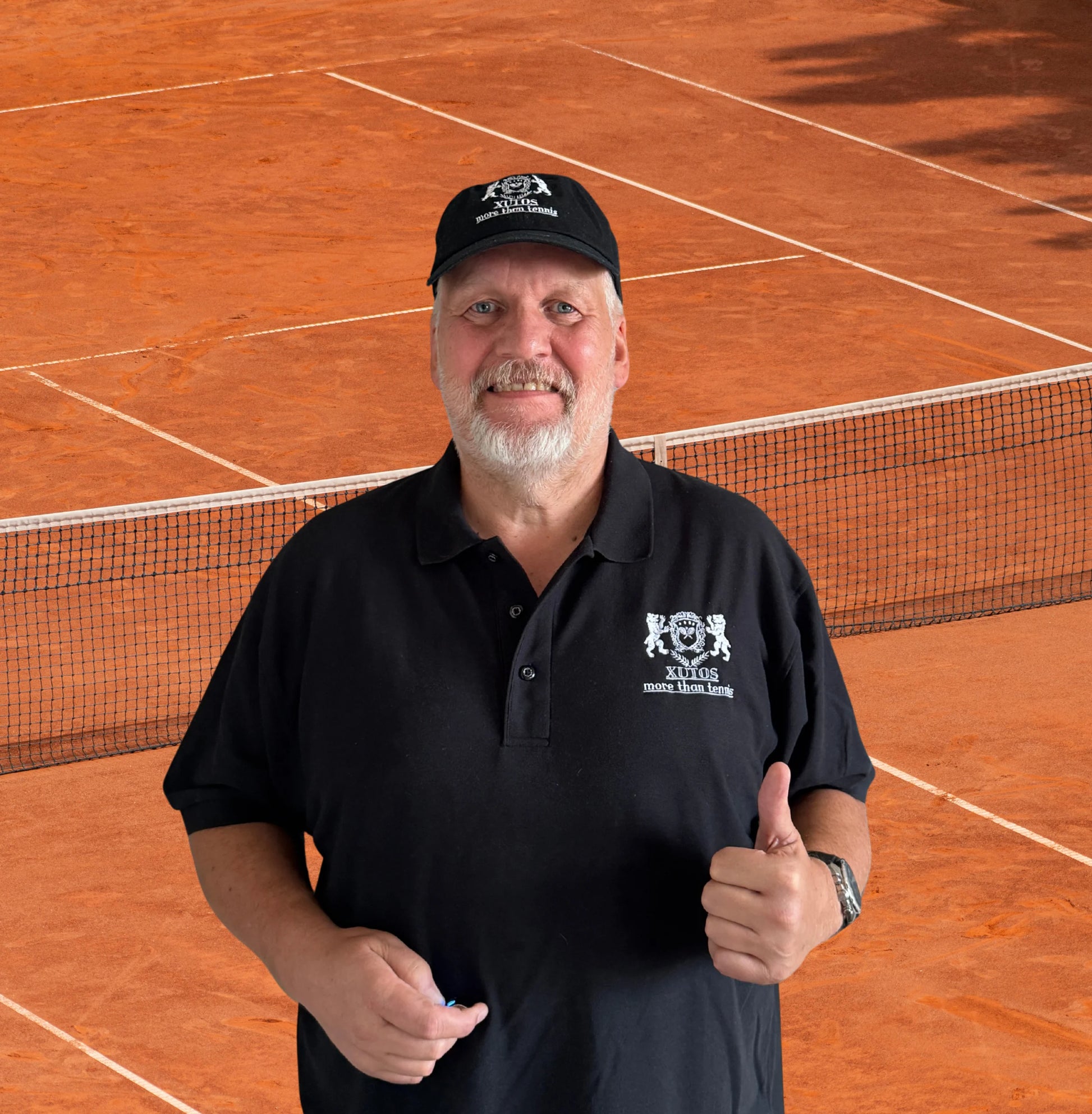 Xutos Polo A smiling man with a white beard wearing a black polo shirt and cap with a white logo, standing on a clay tennis court and giving a thumbs-up.