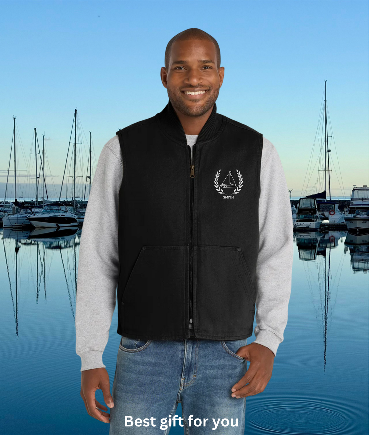 sailing vest A man wearing a black zip-up vest over a light gray long-sleeve shirt and blue jeans, standing in front of a marina background with boats on calm water. The vest has a white sailboat emblem on the left chest with the text 'SAILORS UNITED'.