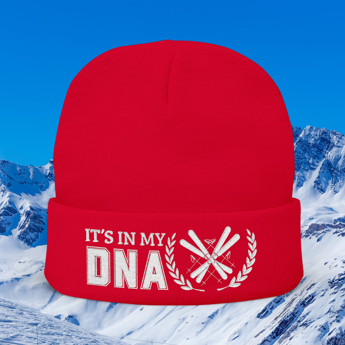 Ski Beanie A bright red knit beanie with white embroidered text reading 'IT'S IN MY DNA' and a crossed-ski graphic, shown against a snowy mountain and blue sky background.