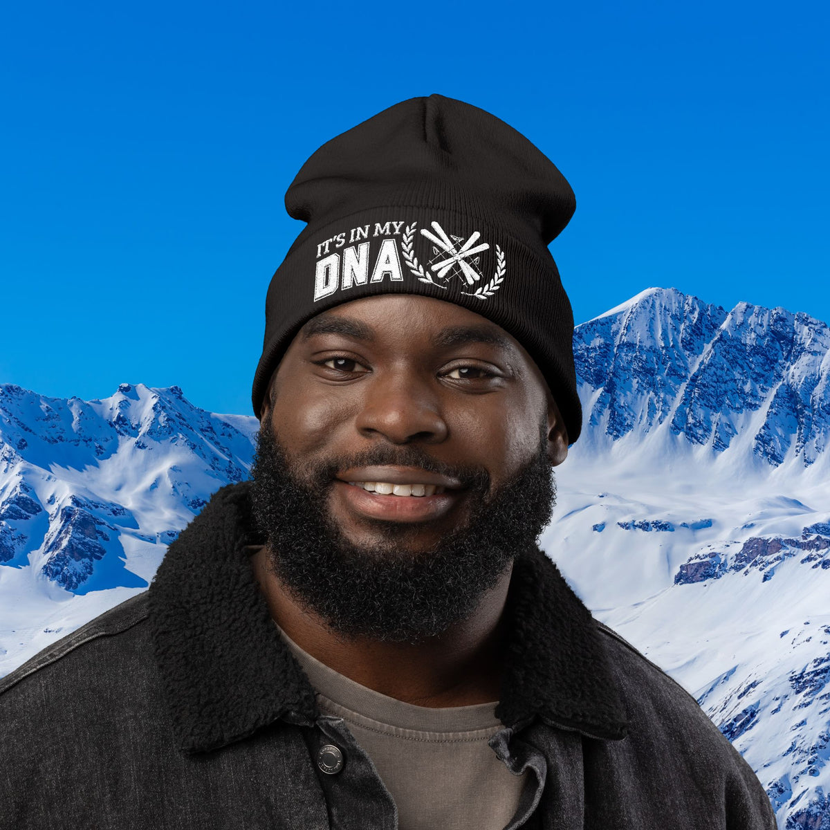 Ski Beanie A smiling man wearing a black beanie with the text 'IT'S IN MY DNA' and a mountain backdrop.