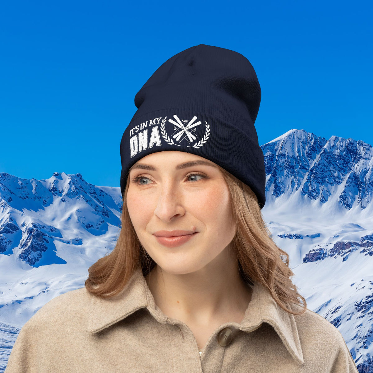 Ski Beanie A woman wearing a black beanie with white embroidered text reading 'IT'S IN MY DNA' and a snowflake motif, set against a snowy mountain and blue sky background.