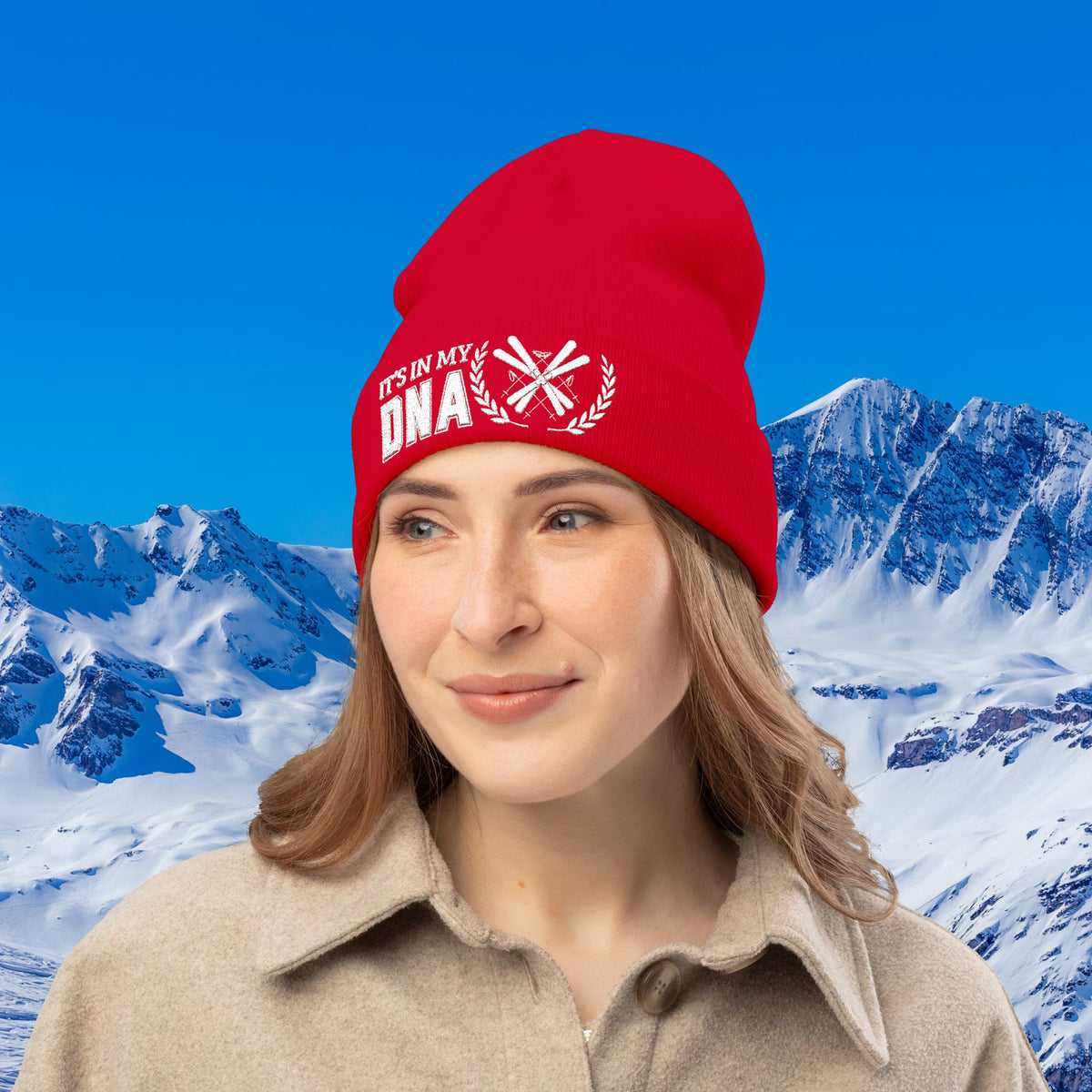 Ski Beanie A woman wearing a bright red beanie with white embroidered text reading 'IT'S IN MY DNA' and a snowflake motif, set against a snowy mountain and blue sky background.