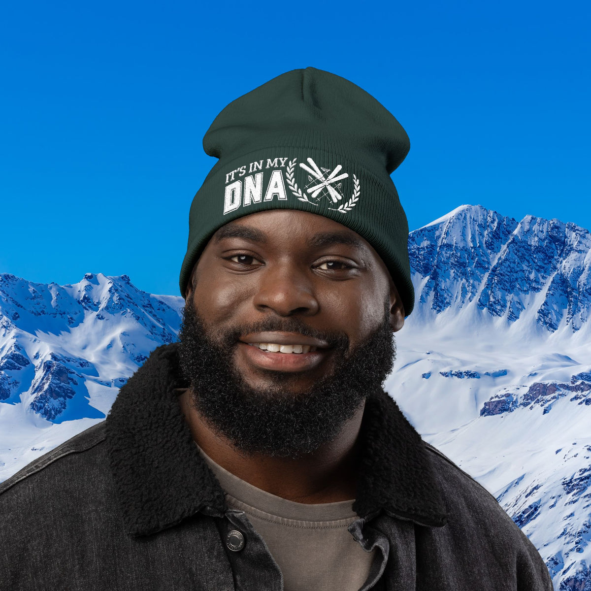 Ski Beanie A smiling man wearing a dark green beanie with the text 'IT'S IN MY DNA' and a mountain backdrop.