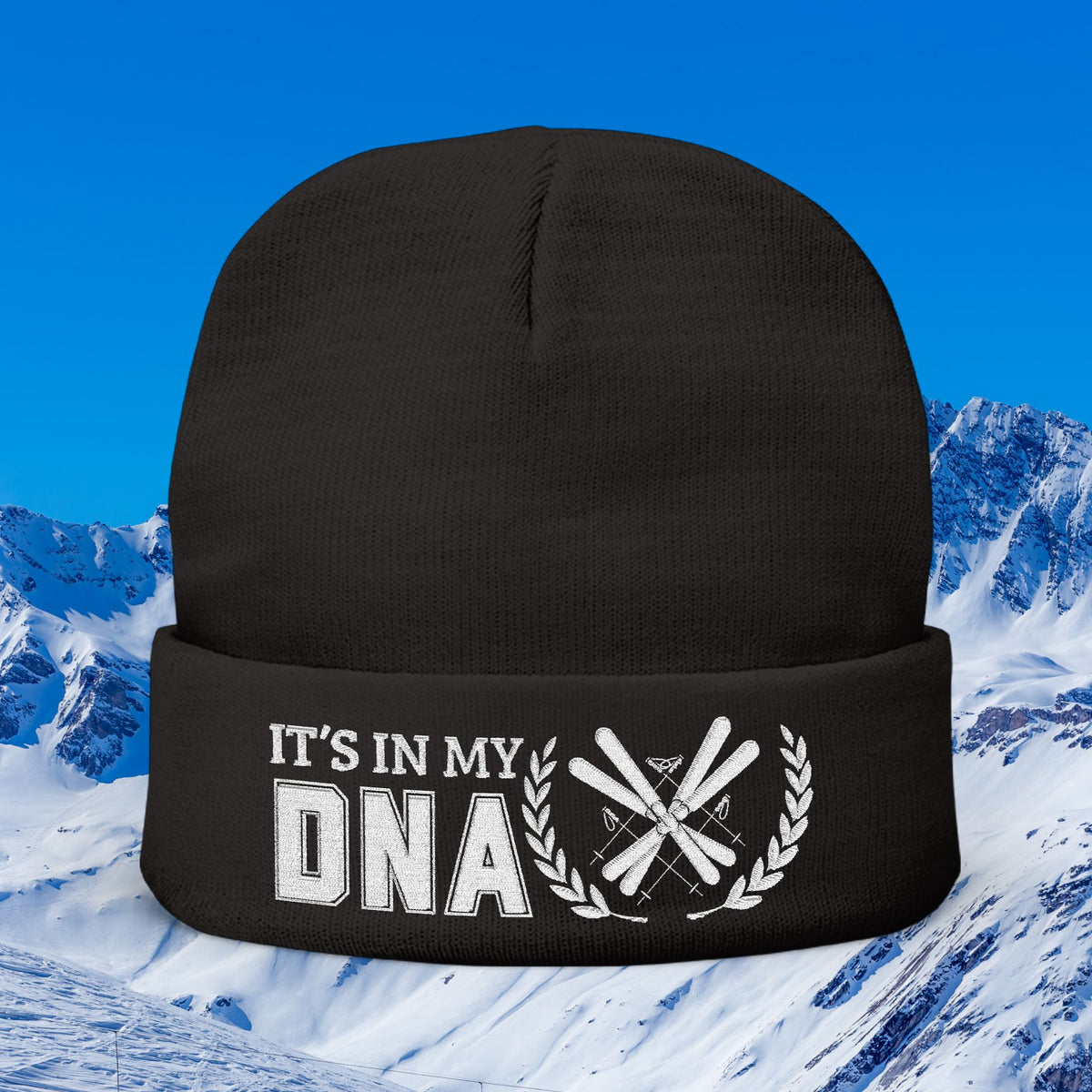 Ski Beanie A black knit beanie with a white graphic on the cuff that reads 'IT'S IN MY DNA' alongside crossed skis and a laurel wreath, shown against a snowy mountain and blue sky background.