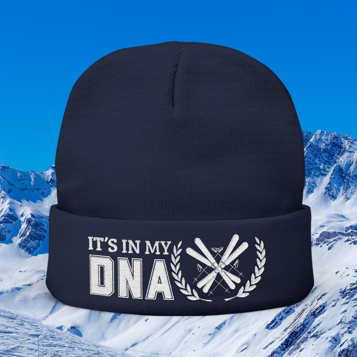 Ski Beanie A dark navy blue knit beanie with a white graphic print on the cuff that reads 'IT'S IN MY DNA' alongside crossed skis and a laurel wreath, set against a snowy mountain background under a blue sky.