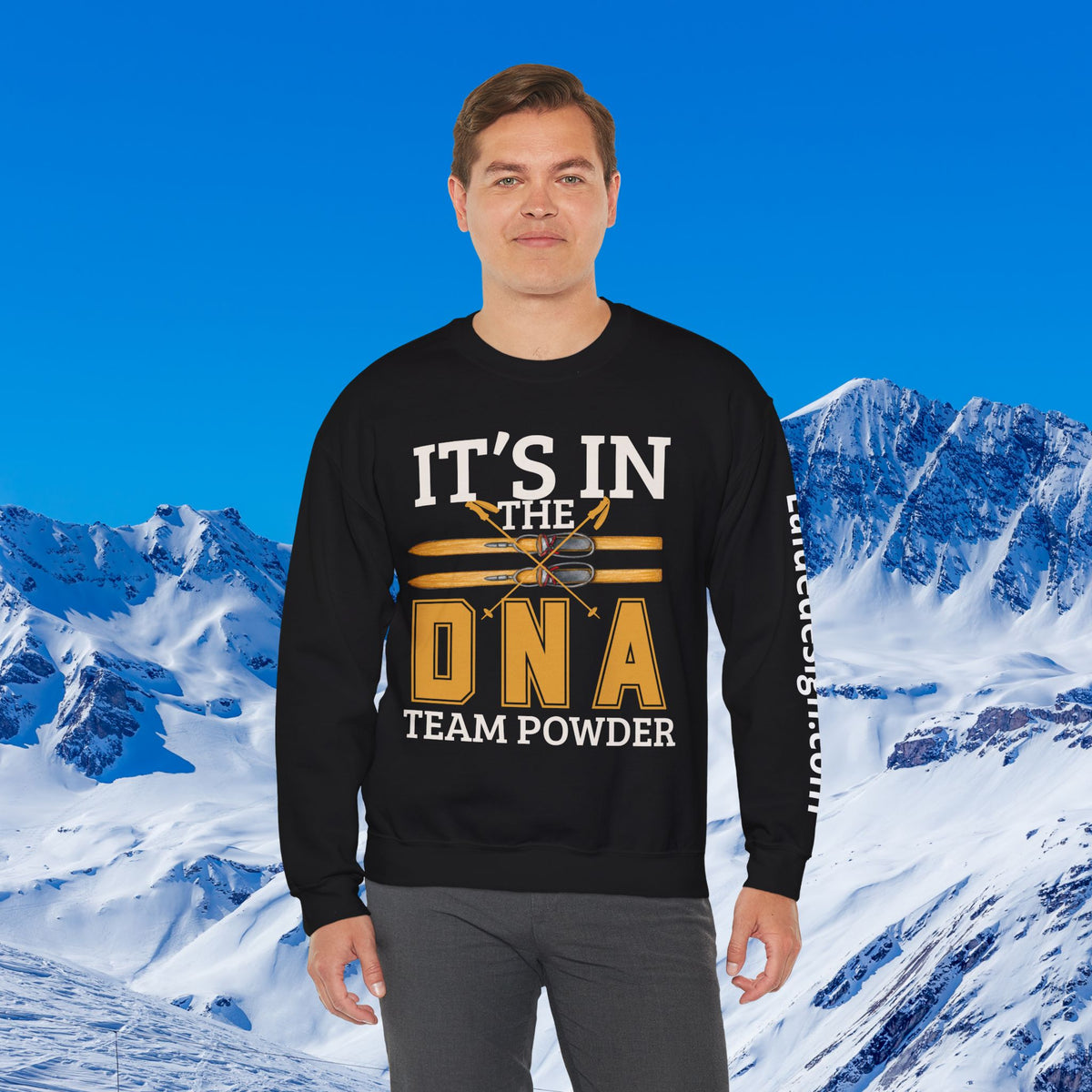 Ski Sweatshirt A man wearing a black crewneck sweatshirt with a yellow and white graphic that reads 'IT'S IN THE DNA TEAM POWDER' stands in front of a snowy mountain background under a blue sky.