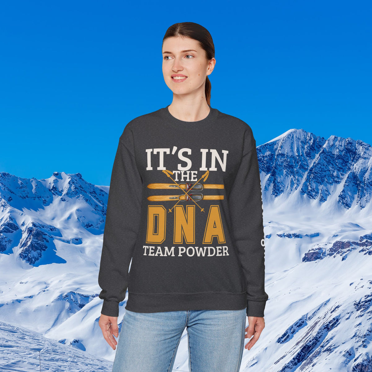 Ski Sweatshirt A woman modeling a dark gray crewneck sweatshirt with a graphic that reads 'IT'S IN THE DNA TEAM POWDER' in white and yellow, set against a snowy mountain background.