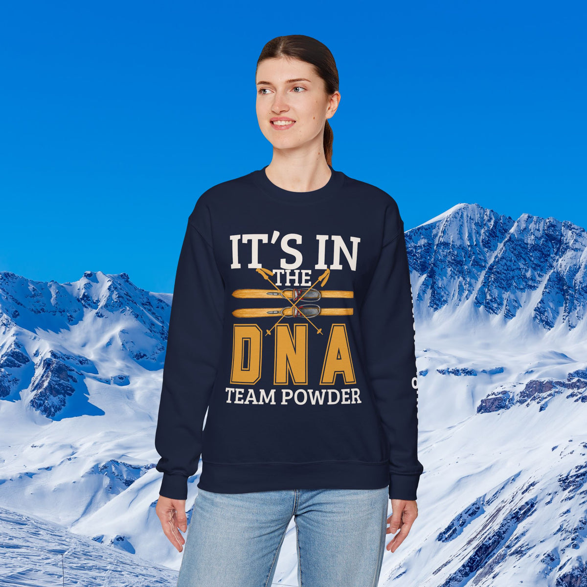 Ski Sweatshirt A woman modeling a navy blue crewneck sweatshirt with a graphic that reads 'IT'S IN THE DNA TEAM POWDER' in white and gold, set against a snowy mountain background.