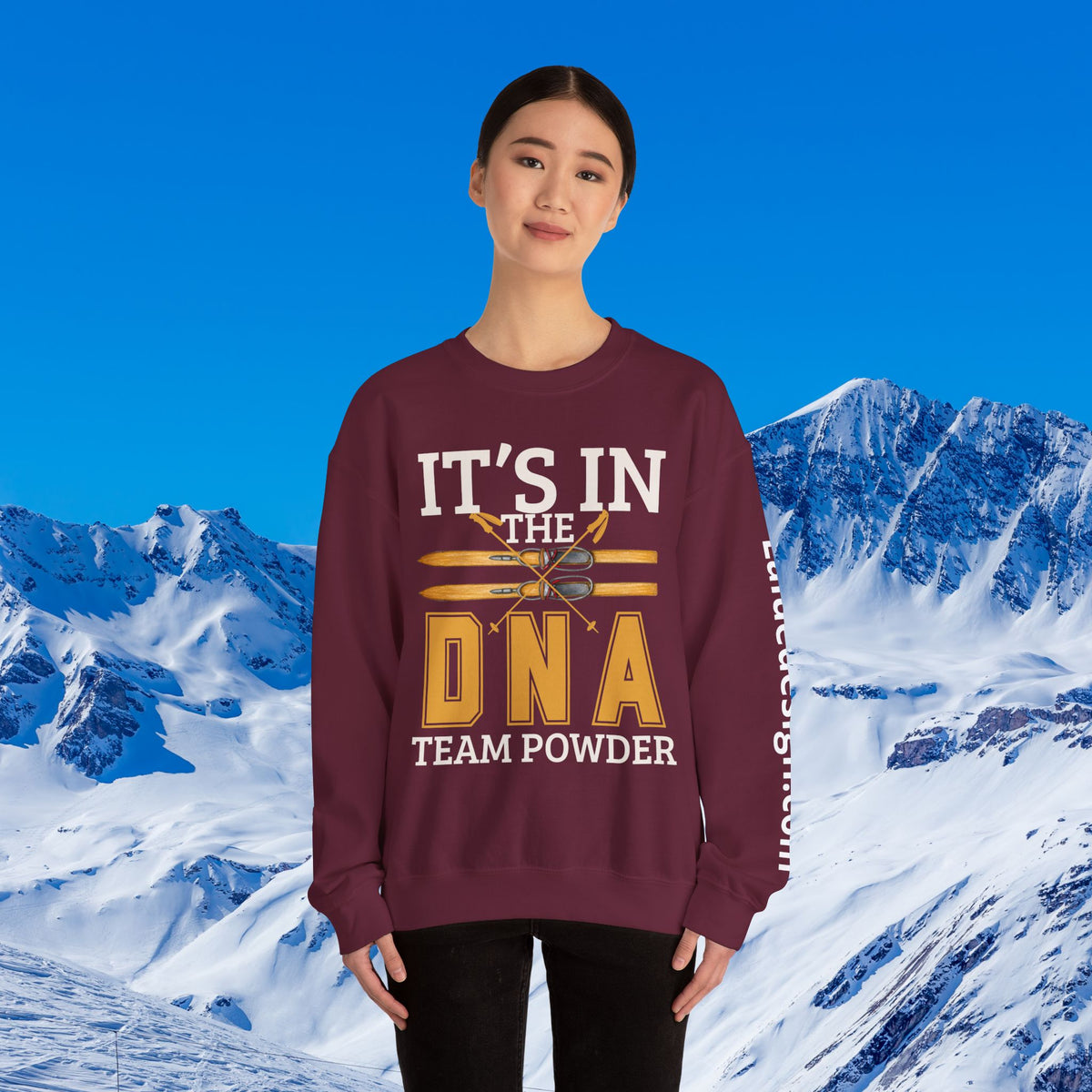 Ski Sweatshirt A woman modeling a maroon crewneck sweatshirt with a graphic that reads 'IT'S IN THE DNA TEAM POWDER' in white and yellow, set against a snowy mountain background.