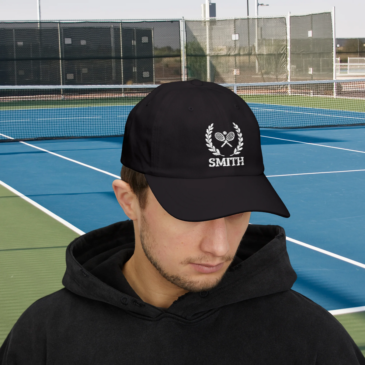 Tennis Cap A man wearing a black baseball cap with a white embroidered tennis-themed logo and the name 'SMITH' on the front, standing on a blue and green tennis court.
