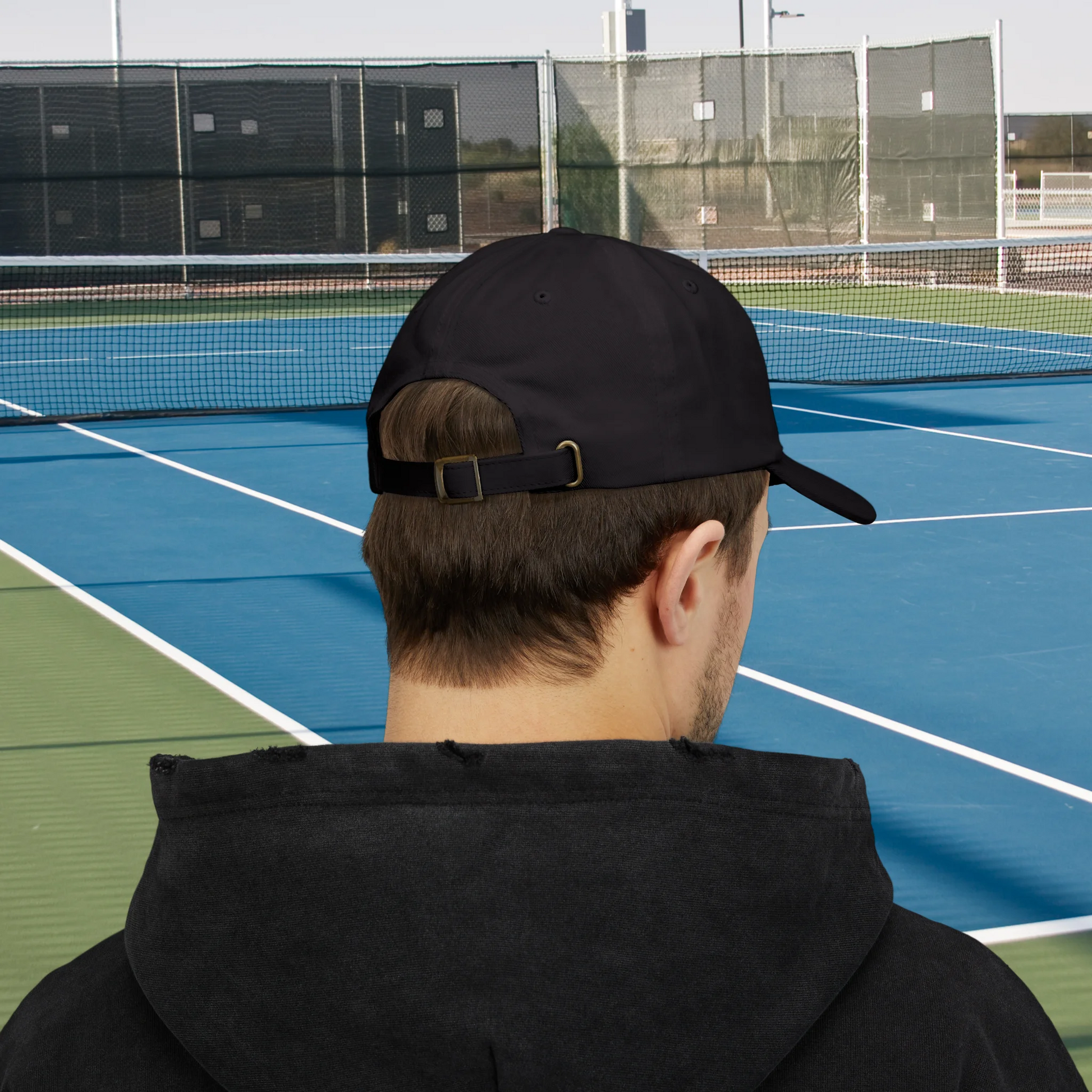Tennis Cap A man viewed from behind wearing a black baseball cap and a black hoodie, standing on a blue and green tennis court with a net and fencing in the background.