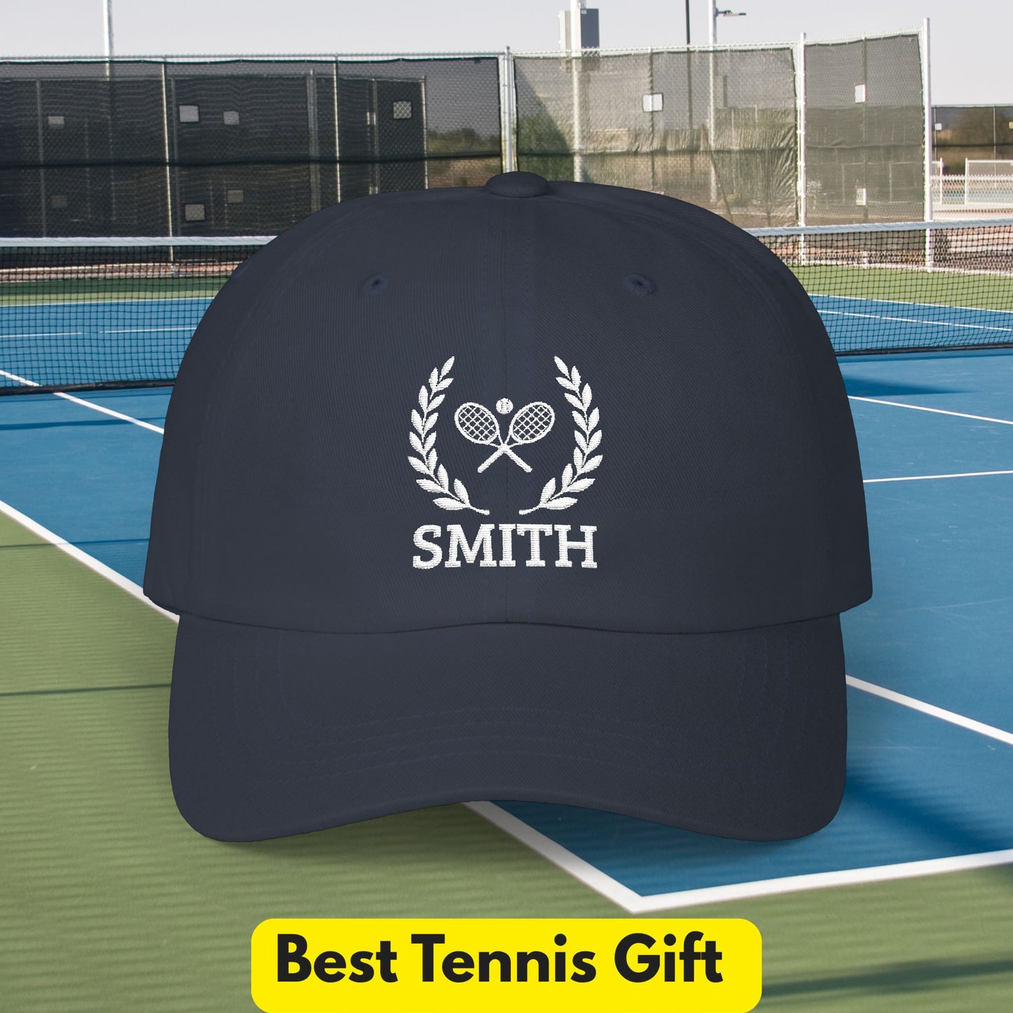 Tennis Cap A dark navy baseball cap with a white embroidered tennis-themed graphic featuring crossed rackets, a laurel wreath, and the name 'SMITH', shown on a tennis court with a yellow 'Best Tennis Gift' banner at the bottom.