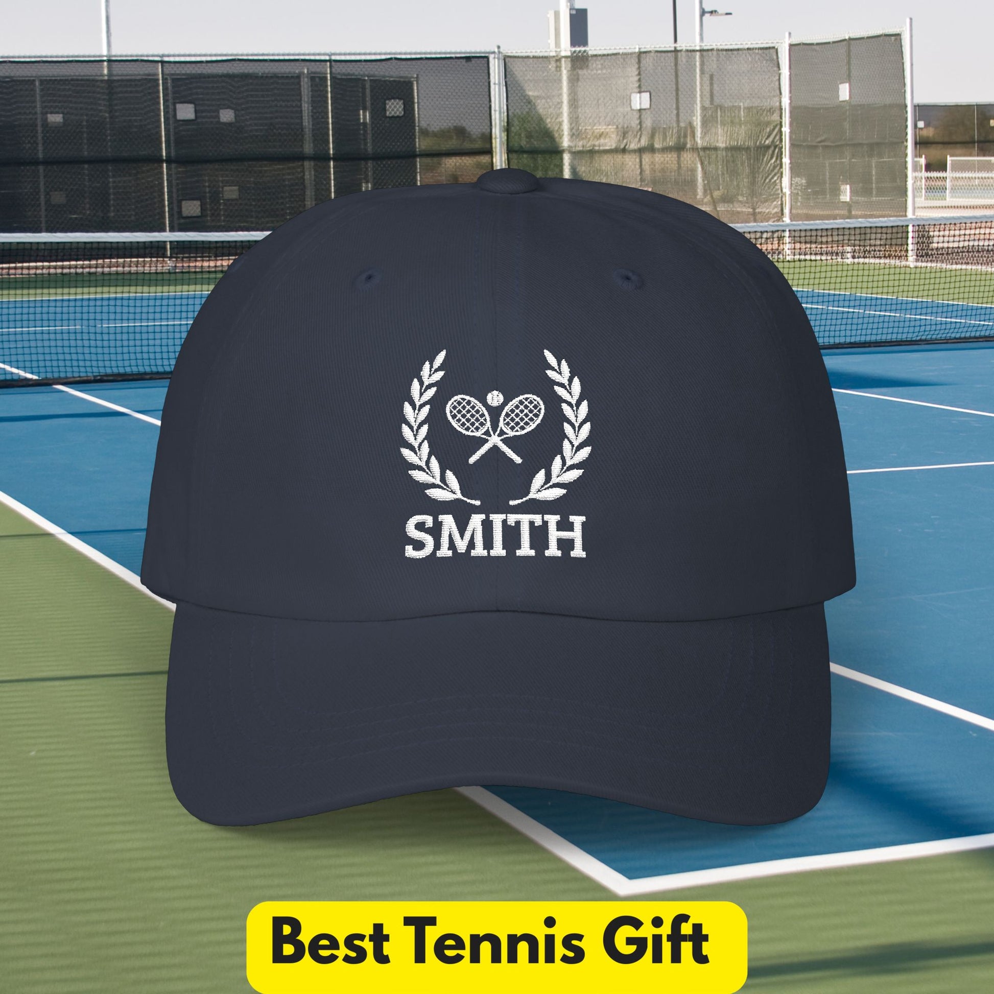 Tennis Cap A dark navy baseball cap with a white embroidered tennis-themed graphic featuring crossed rackets, a laurel wreath, and the name 'SMITH', shown on a tennis court with a yellow 'Best Tennis Gift' banner at the bottom.