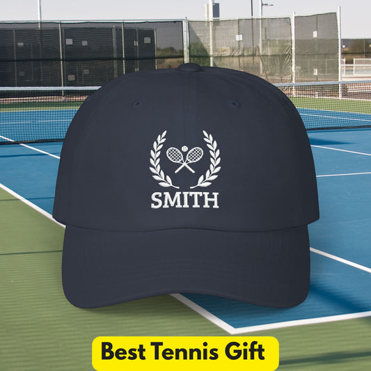 Tennis Cap A dark navy baseball cap with a white embroidered tennis-themed graphic featuring crossed rackets, a laurel wreath, and the name 'SMITH', shown on a tennis court with a yellow 'Best Tennis Gift' banner at the bottom.