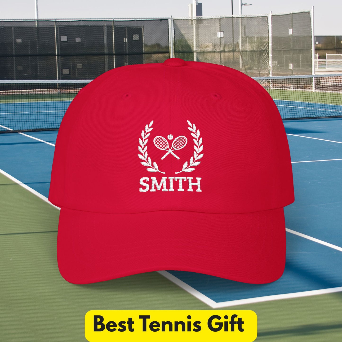 Tennis Cap A red baseball cap with a white embroidered tennis-themed logo and the name 'SMITH' on the front, shown against a tennis court background. A yellow banner at the bottom reads 'Best Tennis Gift'.
