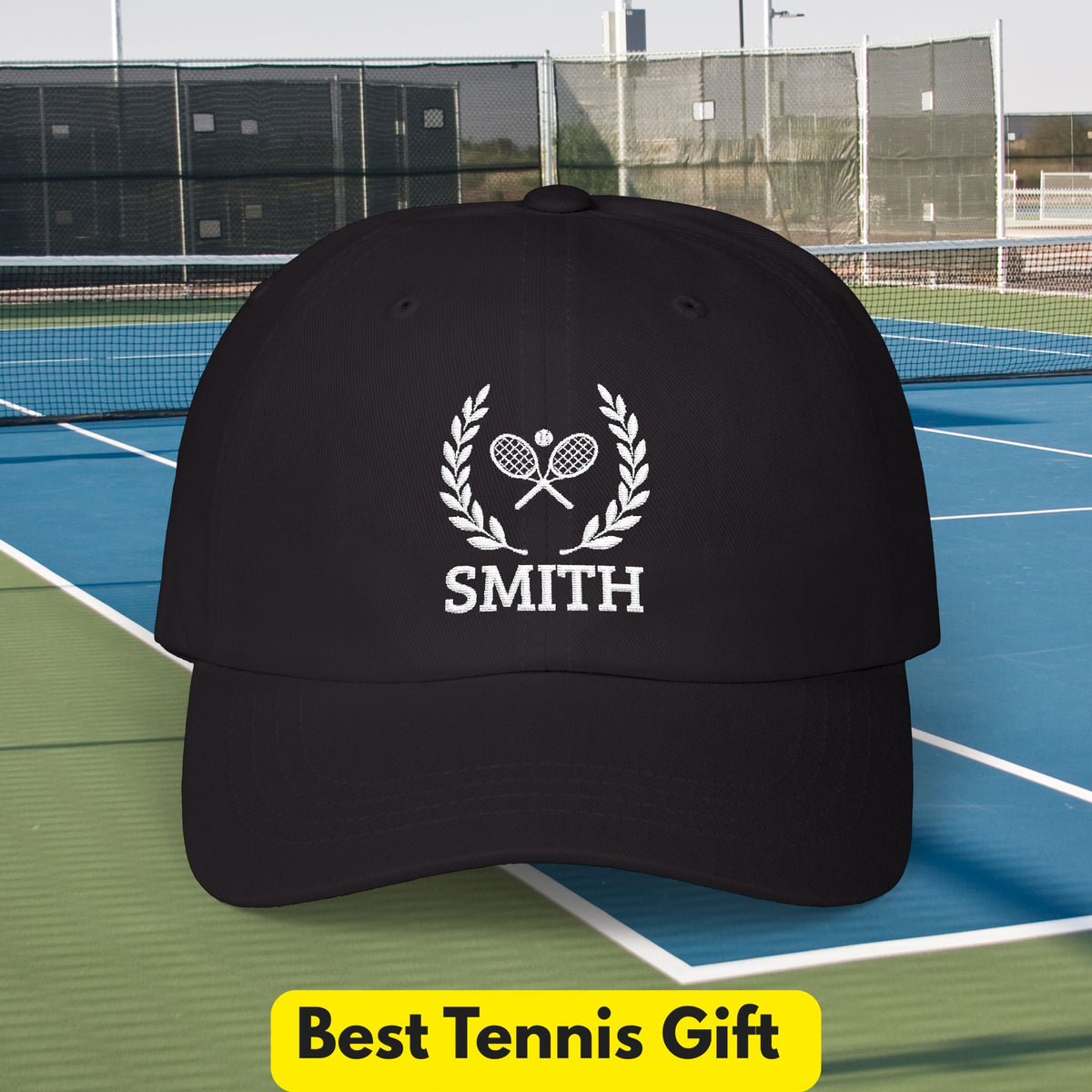 Tennis Cap A black baseball cap with a white embroidered tennis-themed graphic featuring crossed rackets, a laurel wreath, and the name 'SMITH', shown on a tennis court with a yellow 'Best Tennis Gift' banner at the bottom.