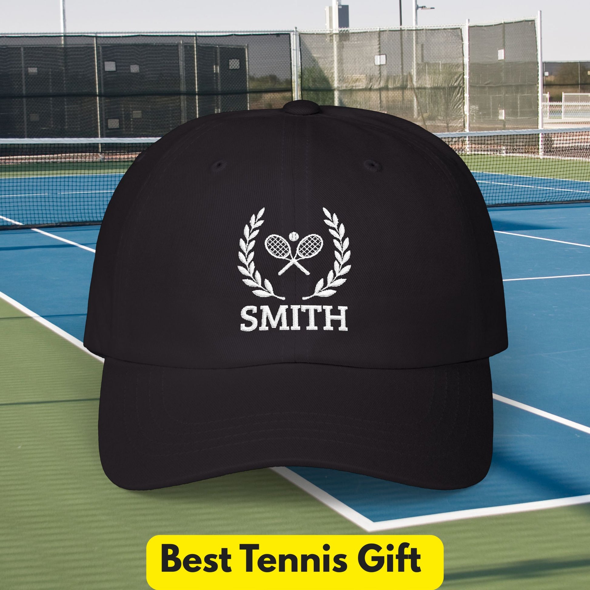 Tennis Cap A black baseball cap with a white embroidered tennis-themed graphic featuring crossed rackets, a laurel wreath, and the name 'SMITH', shown on a tennis court with a yellow 'Best Tennis Gift' banner at the bottom.