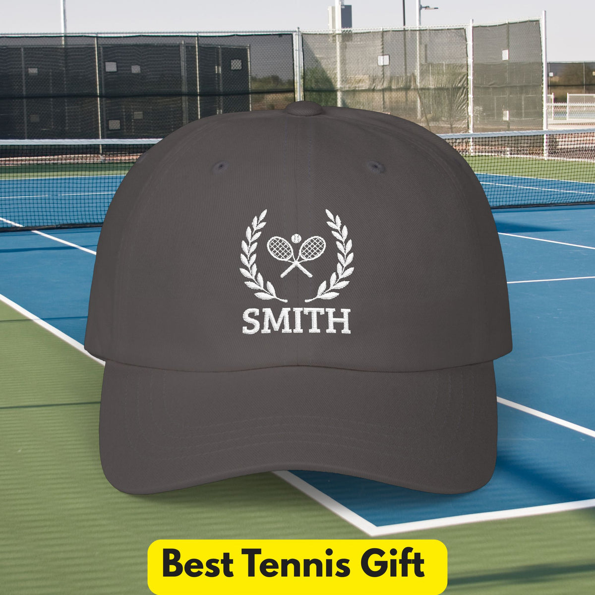 Tennis Cap A dark gray baseball cap with a white tennis-themed graphic featuring crossed rackets, a laurel wreath, and the name 'SMITH' printed on the front. The cap is shown on a blue and green tennis court with a yellow 'Best Tennis Gift' banner at the bottom.