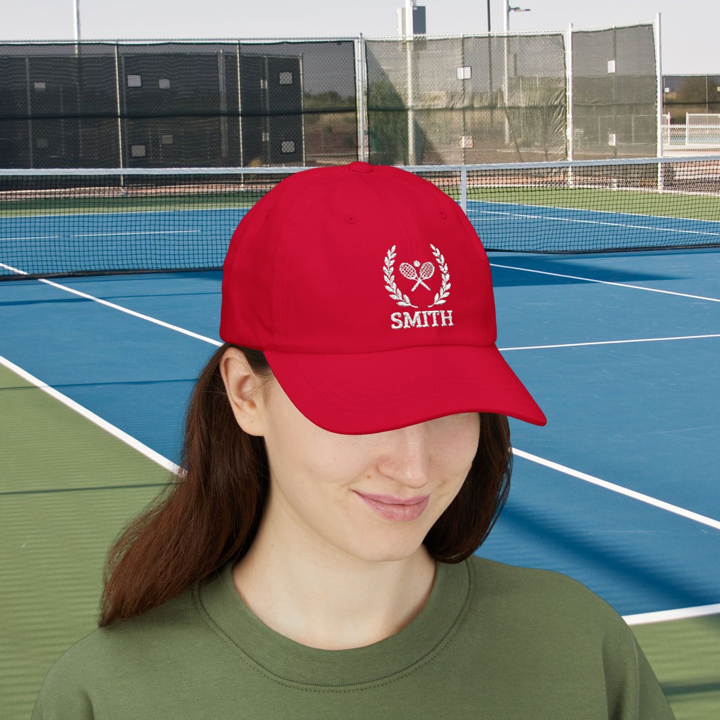 Tennis Cap A woman wearing a red baseball cap with a white embroidered laurel wreath, crossed tennis rackets, and the name 'SMITH' on the front, photographed on a blue and green tennis court.