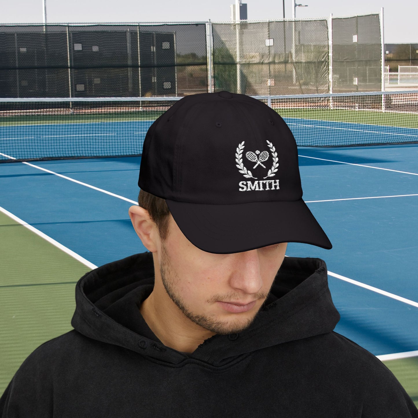 Tennis Cap A man wearing a black baseball cap with a white embroidered tennis-themed logo and the name 'SMITH' on the front, standing on a blue and green tennis court.