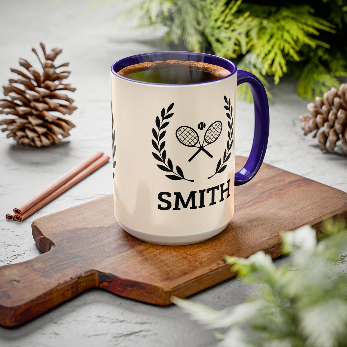 Tennis Coffee Mug A cream-colored ceramic coffee mug with a dark blue rim and handle, featuring a black tennis racket graphic and the name 'SMITH' printed on the front. The mug is filled with coffee and sits on a wooden cutting board surrounded by pinecones and green foliage.