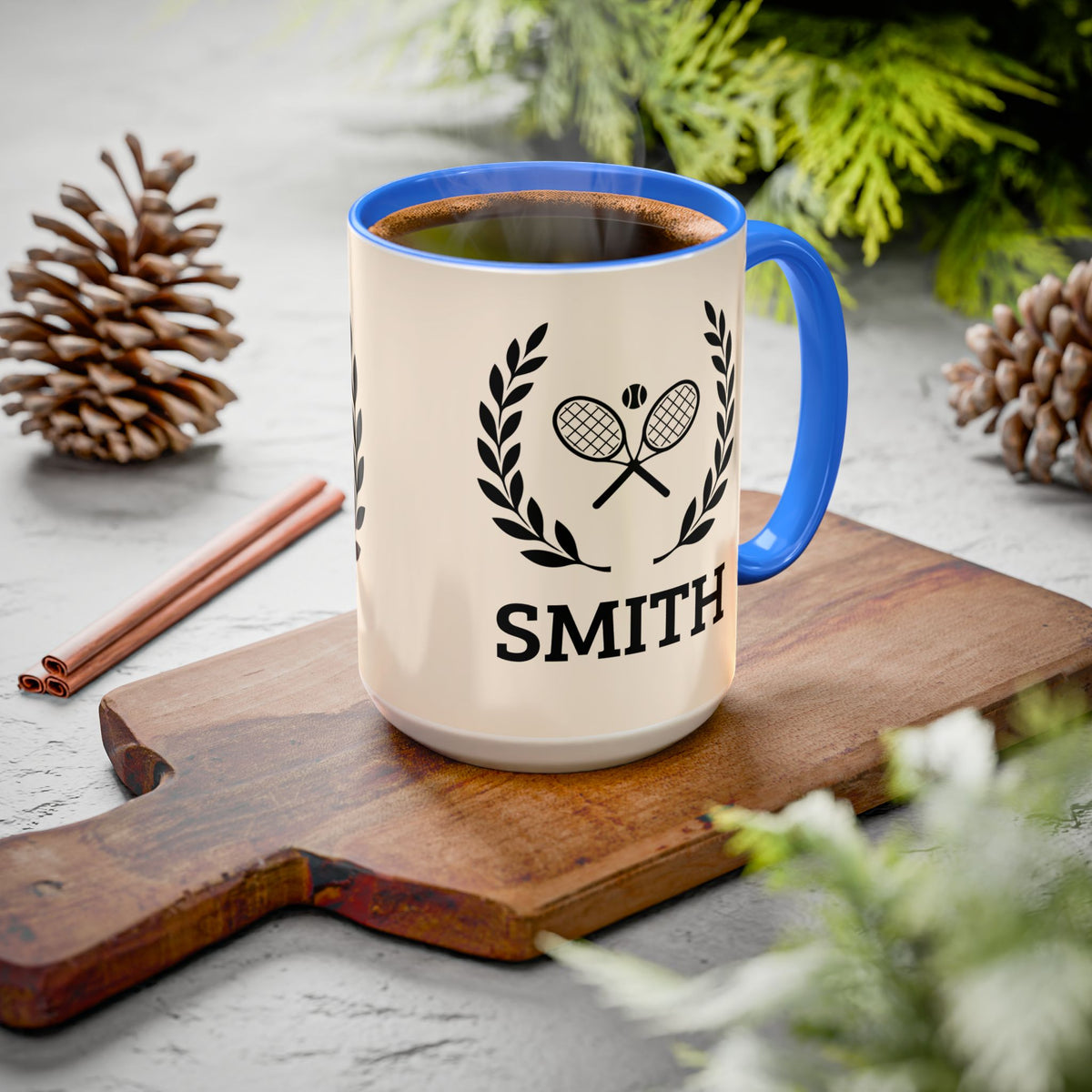 Tennis Coffee Mug A ceramic coffee mug with a cream body, blue rim and handle, and a black tennis racket graphic with the name 'SMITH' below it, styled on a wooden cutting board with pinecones and greenery.