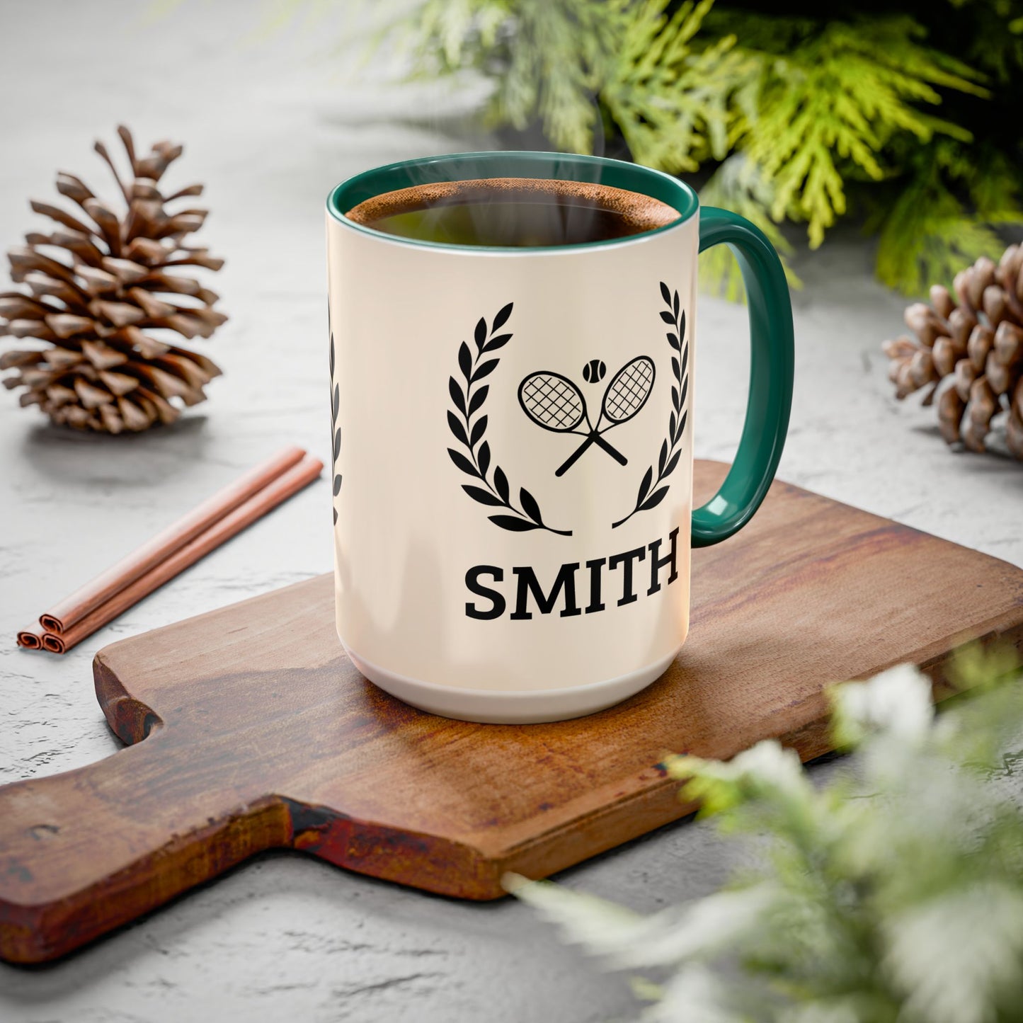 Tennis Coffee Mug A personalized ceramic mug with a green handle and rim, filled with a hot beverage, resting on a wooden cutting board. The mug is cream-colored with a black graphic of crossed tennis rackets under a laurel wreath and the name 'SMITH' printed below.
