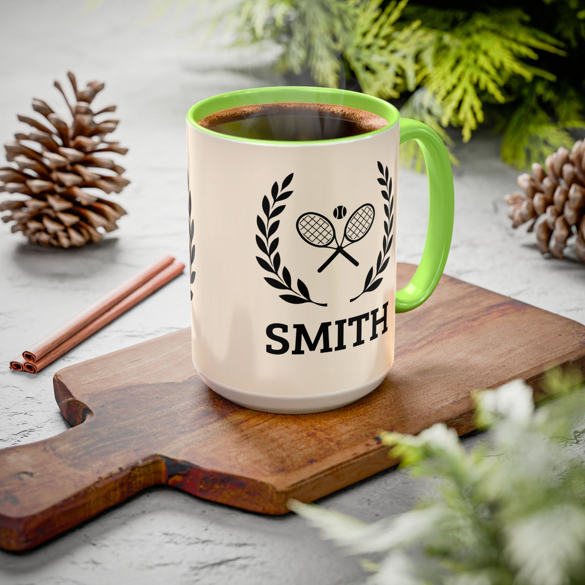Tennis Coffee Mug A ceramic mug filled with a hot beverage sits on a wooden cutting board in a styled setting with pinecones, cinnamon sticks, and greenery. The mug is off-white with a bright green handle and interior rim, featuring a black graphic of crossed tennis rackets under a laurel wreath and the name 'SMITH' printed below.