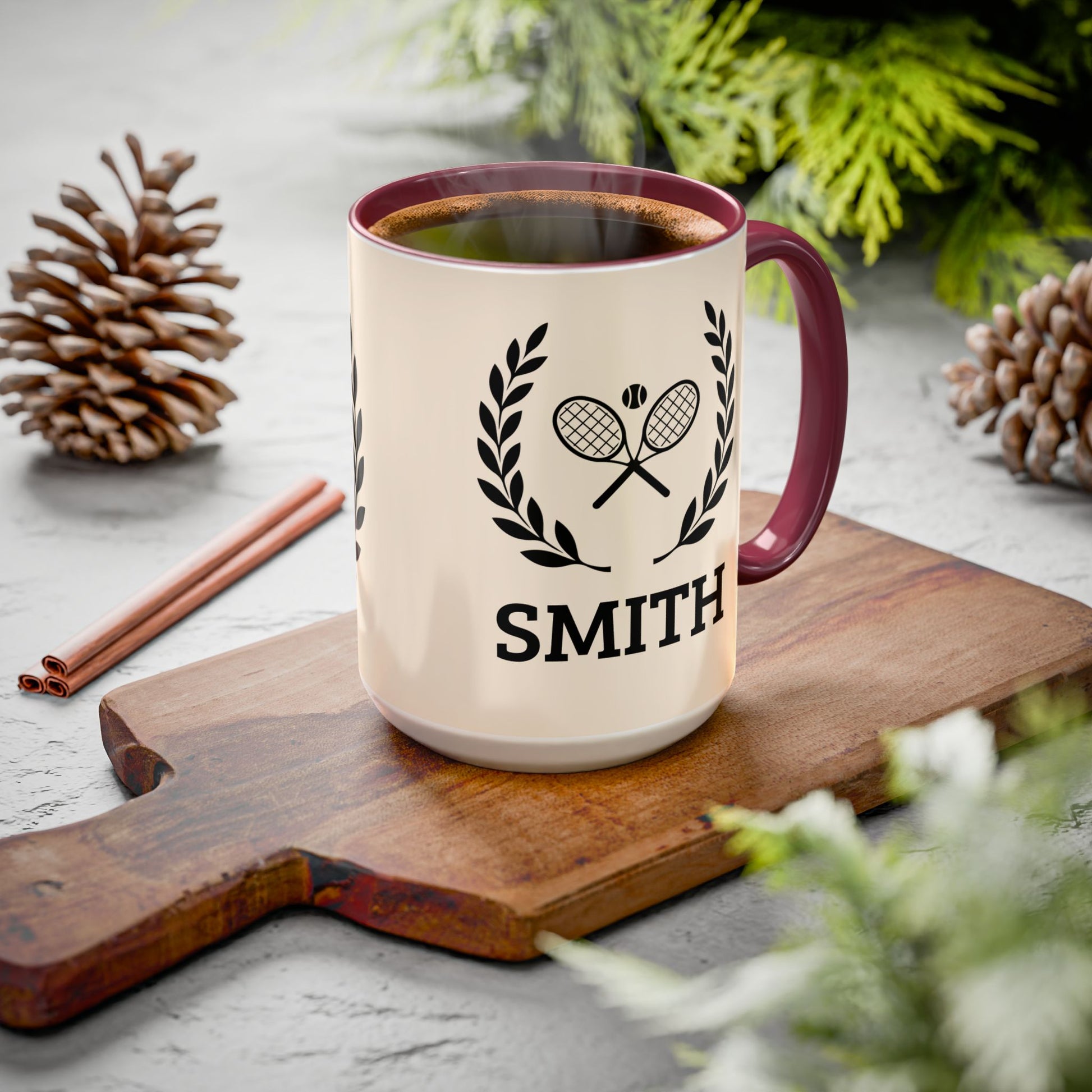 Tennis Coffee Mug A cream-colored ceramic mug with a dark red handle and rim, filled with a hot beverage. The mug features a black graphic of two crossed tennis rackets enclosed in a laurel wreath, with the name 'SMITH' printed below.