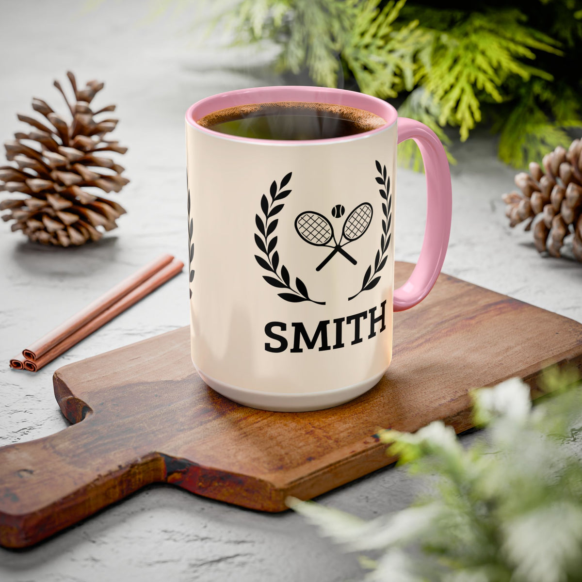Tennis Coffee Mug A personalized ceramic coffee mug with a pink handle and rim, filled with black coffee. The mug is cream-colored with a black graphic of two crossed tennis rackets under a laurel wreath and the name 'SMITH' printed below.