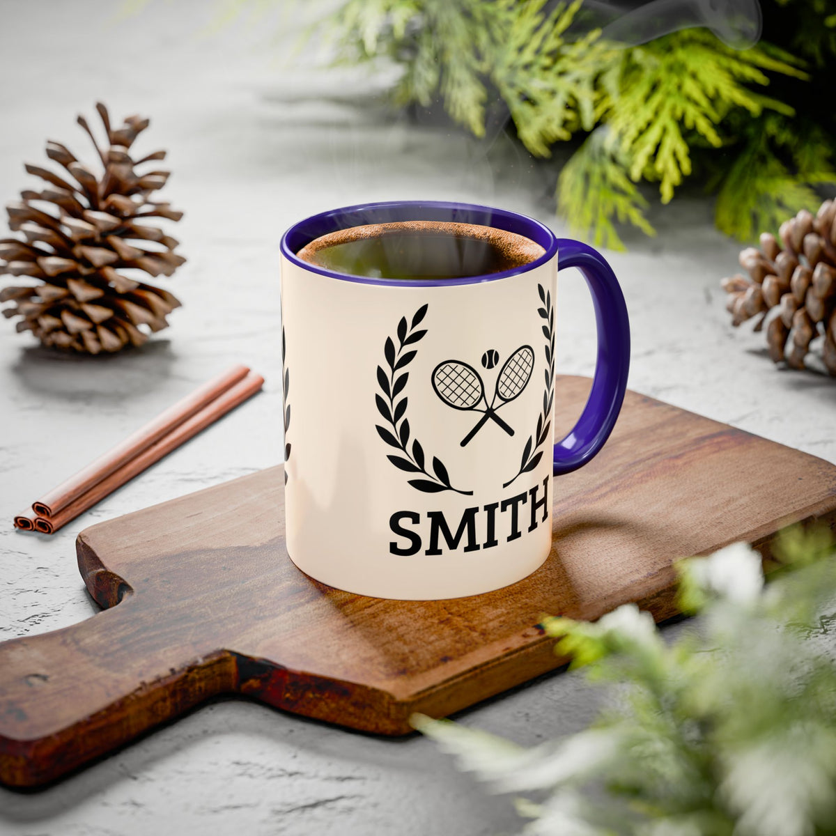 Tennis Coffee Mug A white ceramic coffee mug with a dark purple handle and rim, featuring a black tennis racket and laurel wreath graphic above the name 'SMITH'. The mug is filled with hot coffee and sits on a wooden cutting board surrounded by pinecones, cinnamon sticks, and green foliage.