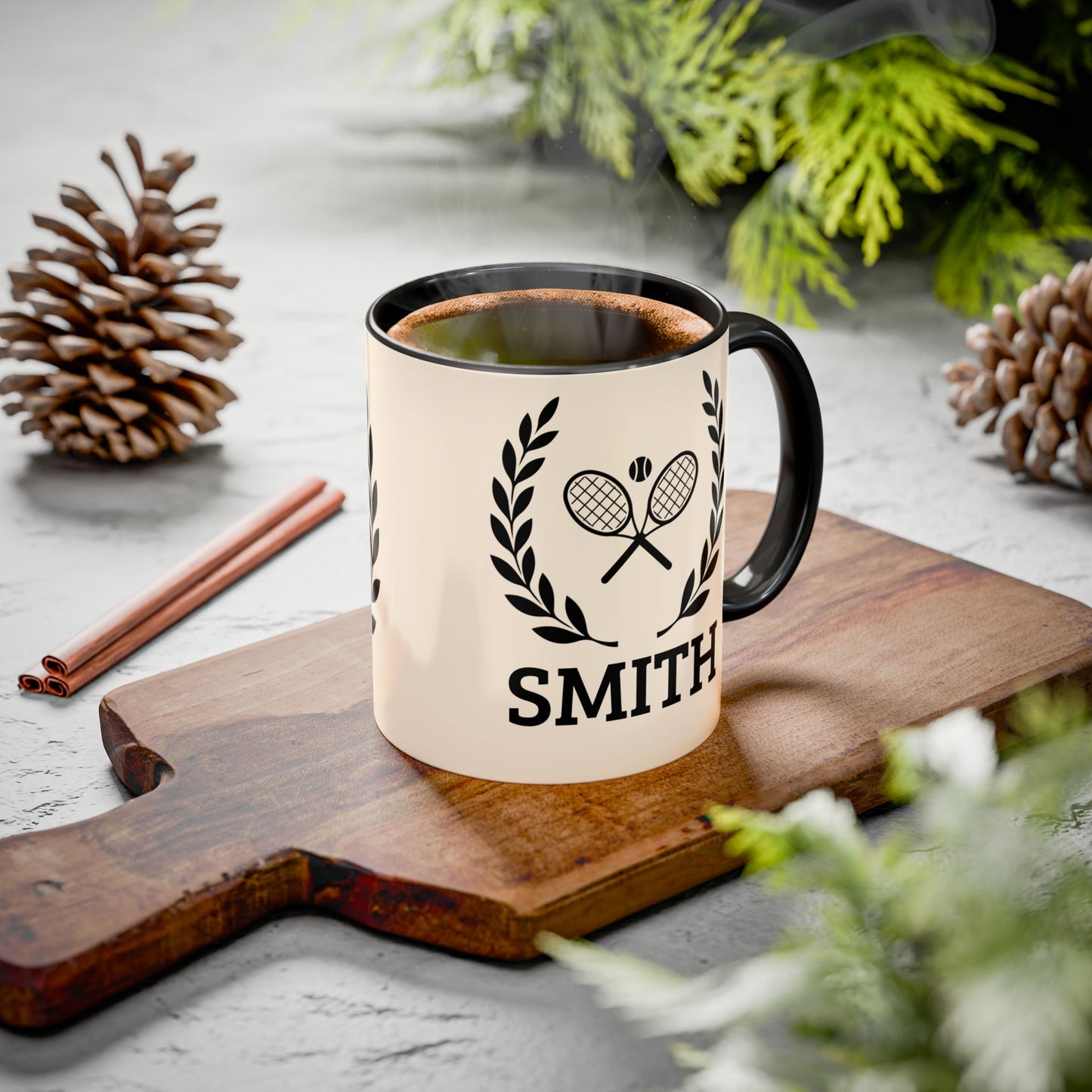 Tennis Coffee Mug A cream-colored ceramic mug with a black handle and rim, featuring a black tennis racket and laurel wreath graphic above the name 'SMITH'. The mug is filled with a hot beverage and sits on a wooden cutting board surrounded by pinecones, cinnamon sticks, and green foliage on a gray stone surface.