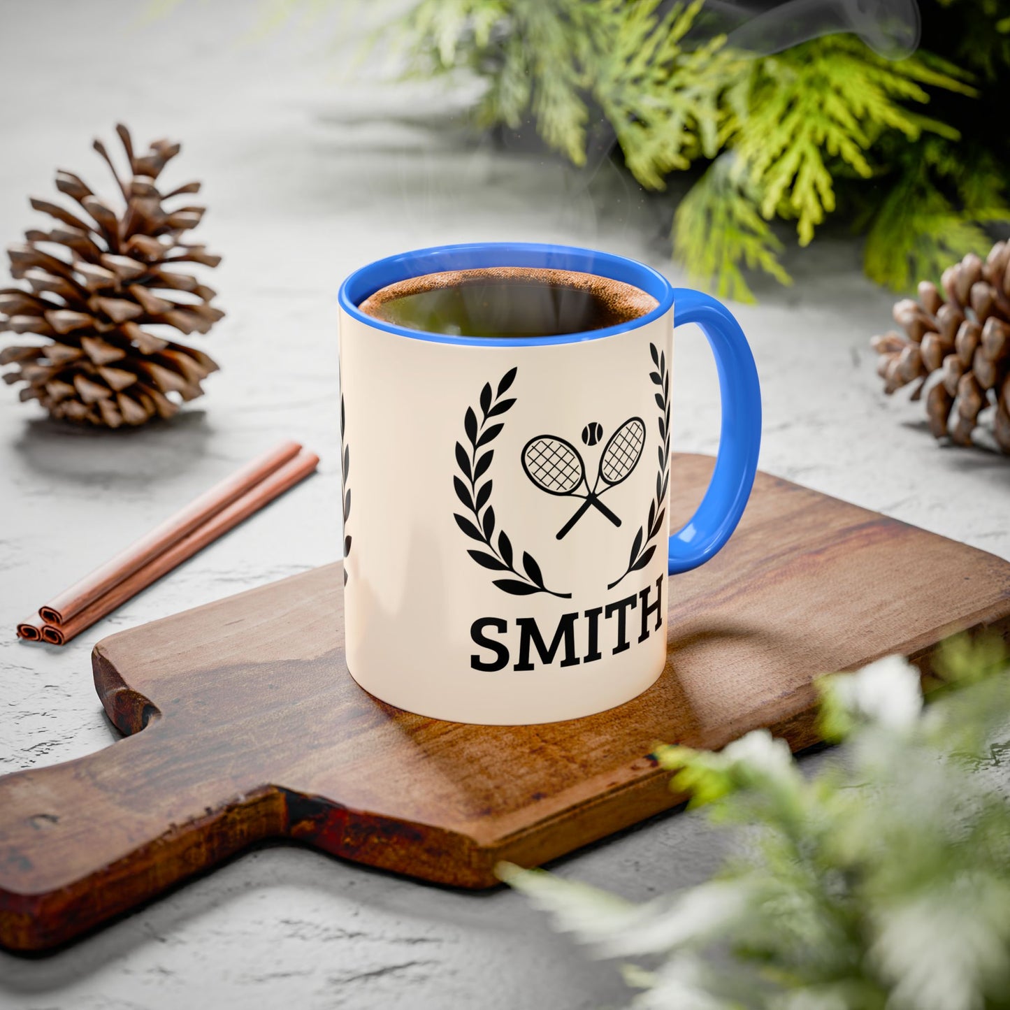 Tennis Coffee Mug A ceramic mug with a blue handle and rim sits on a wooden cutting board, filled with a hot beverage. The mug is cream-colored with a black graphic of crossed tennis rackets inside a laurel wreath and the name 'SMITH' printed below.