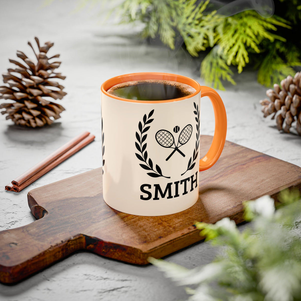 Tennis Coffee Mug A personalized ceramic coffee mug with an orange handle and rim, featuring a black tennis racket and laurel wreath graphic above the name 'SMITH'. The mug sits on a wooden cutting board surrounded by pinecones, cinnamon sticks, and green foliage on a gray stone surface.