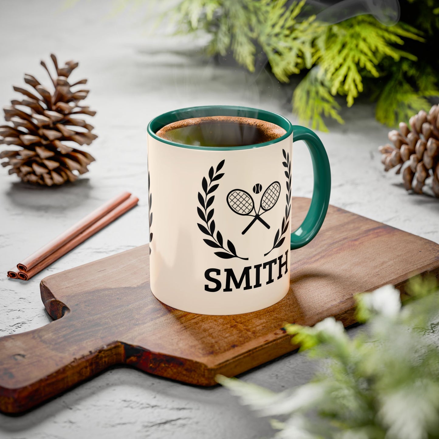 Tennis Coffee Mug A ceramic mug with a cream body, dark green handle and rim, featuring a black tennis racket and laurel wreath graphic above the name 'SMITH'. The mug sits on a wooden cutting board surrounded by pinecones, cinnamon sticks, and green foliage on a light gray surface.