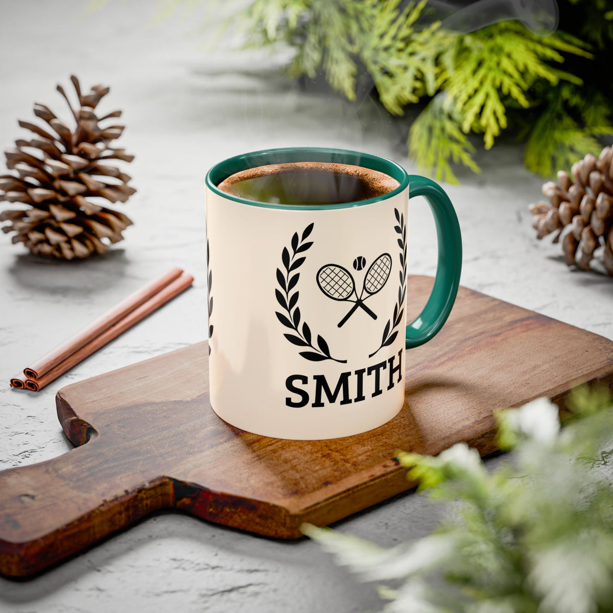 Tennis Coffee Mug A ceramic mug with a cream body, dark green handle and rim, featuring a black tennis racket and laurel wreath graphic above the name 'SMITH'. The mug sits on a wooden cutting board surrounded by pinecones, cinnamon sticks, and green foliage on a light gray surface.
